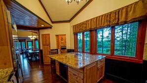 Una cocina amplia con una isla grande, armarios de madera y vistas a un bosque a través de ventanales.