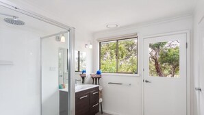 Ein Badezimmer mit einer Duschkabine aus Glas, einem dunklen Holzwaschtisch, einem Fenster mit Blick auf Bäume und einer weißen Tür.