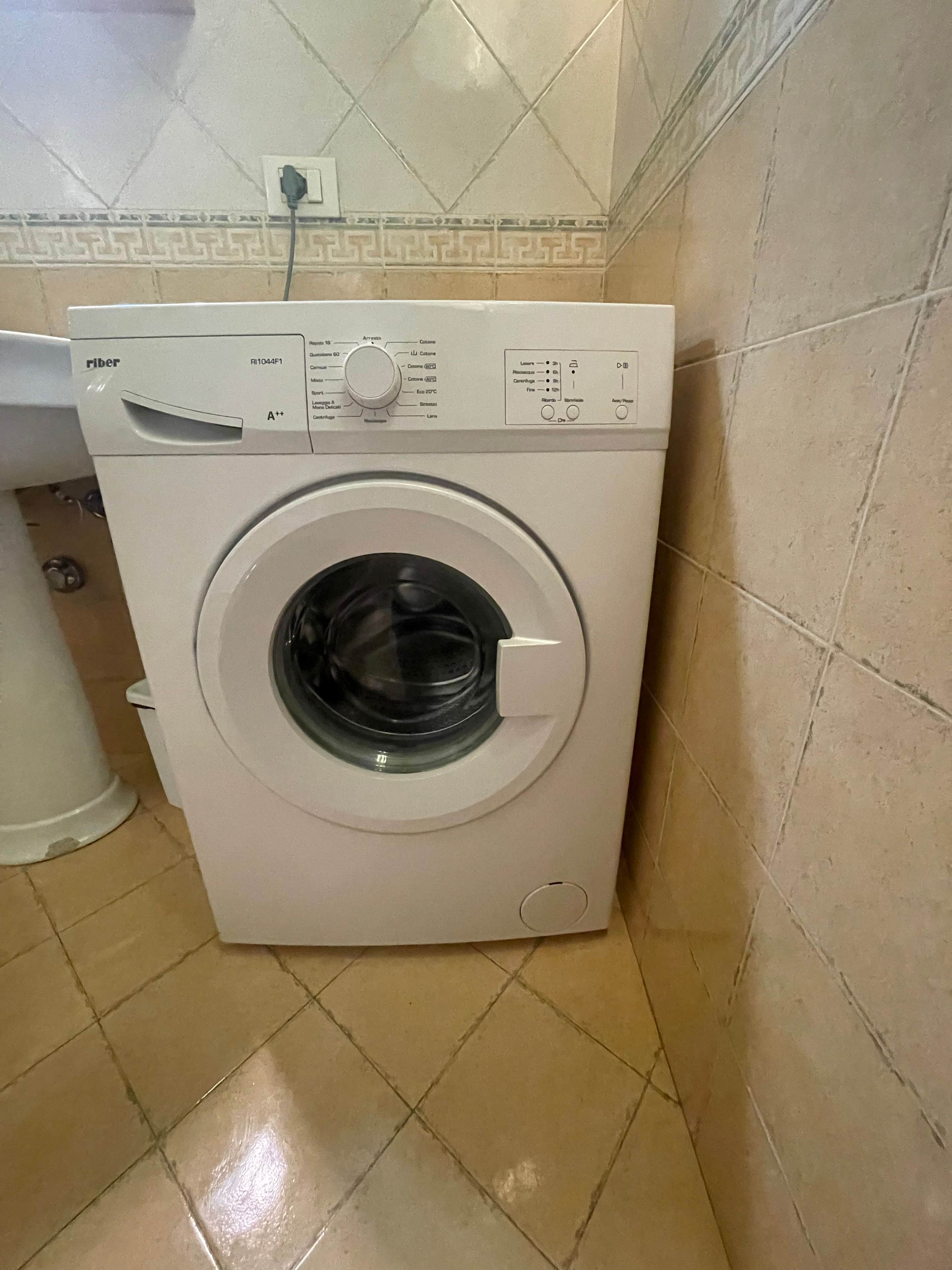 A front-loading washing machine with a control panel, located in a corner of a room with tiled flooring.