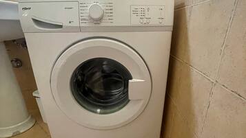 A front-loading washing machine with a control panel, located in a corner of a room with tiled flooring.