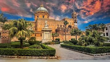 A historic building with a large dome and bell tower, surrounded by well-manicured gardens and palm trees.