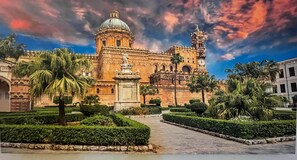 A historic building with a large dome and bell tower, surrounded by well-manicured gardens and palm trees.