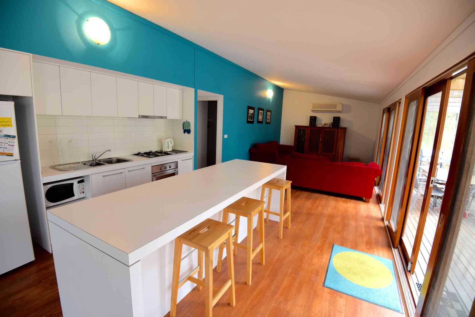 A compact kitchen with white cabinets, a central island with stools, a red sofa in the living area, and a large window.