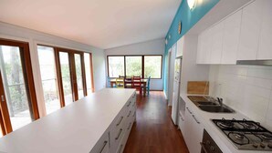 A modern kitchen with a central island, white cabinetry, a gas stove, and a dining area with colorful chairs. - Three Bedroom Bushland House (Anglesea)