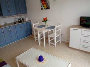 A small kitchen with blue cabinets, a white dining table with chairs, a small white cabinet, and a television on a wooden stand. - apartment at the beach of Getares with roomsbikeanddive (Algeciras)