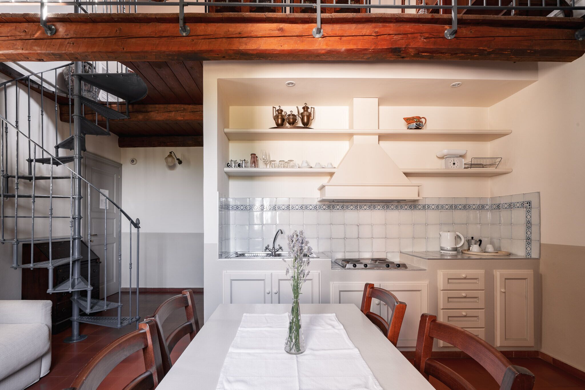 A dining area with a white table, chairs, and a vase of flowers. A spiral staircase is visible to the left. The kitchen area is in the background with white cabinets and a tiled backsplash.