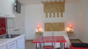 Habitació senzilla amb zona de cuina, un llit amb dues taules de nit, taula vermella amb caires i un sofà vermell.