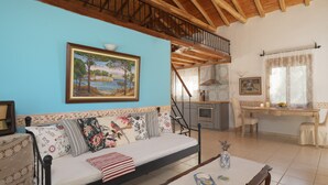 A cozy living room with a wooden ceiling, a sofa with floral cushions, a dining table, and a kitchen area. - Eleni's Charming Cottage, ideal for Couples or Families seeking Natural Living (Skiathos island)