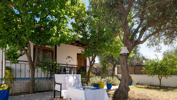 A patio with a table set for dining, surrounded by trees and a building with a balcony. - Eleni's Charming Cottage, ideal for Couples or Families seeking Natural Living (Skiathos island)