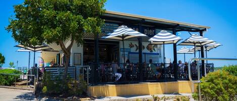 An outdoor restaurant with tables and umbrellas, a building with a sign, and a tree in the foreground.
