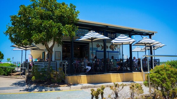 An outdoor restaurant with tables and umbrellas, a building with a sign, and a tree in the foreground. - Dog Friendly House Near Beach (BELLARA)