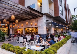 Un área de comedor al aire libre con mesas y sillas, la entrada de un restaurante y la fachada de un edificio con un alero de madera y luces colgantes.