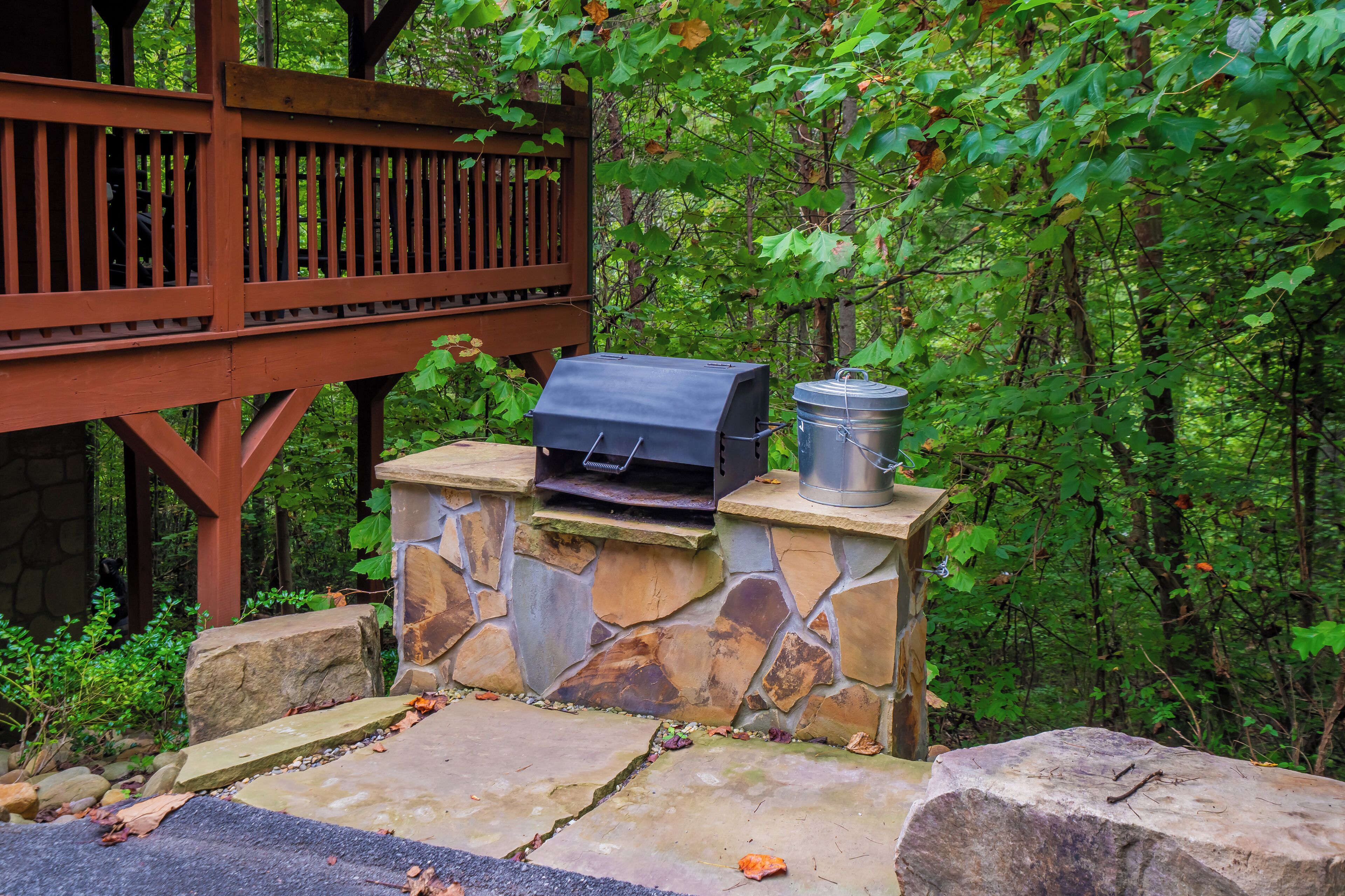 A stone barbecue grill with a metal container, set on a stone platform surrounded by greenery.