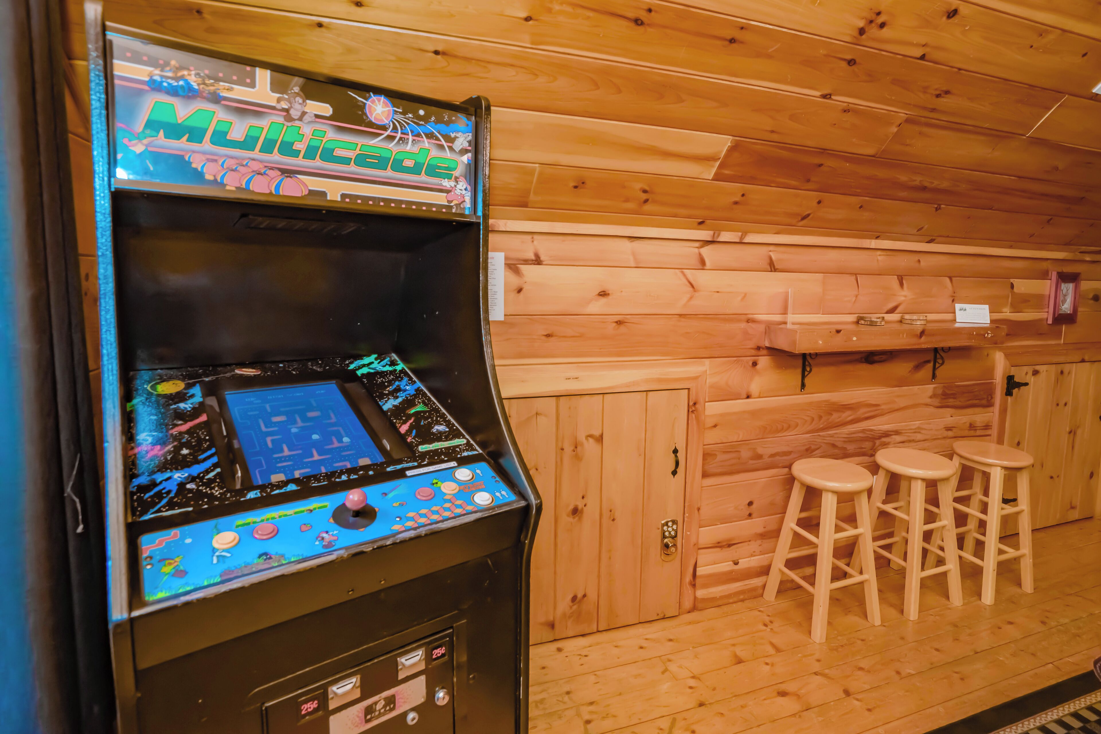 A Multicade arcade machine in a wooden room with three stools.