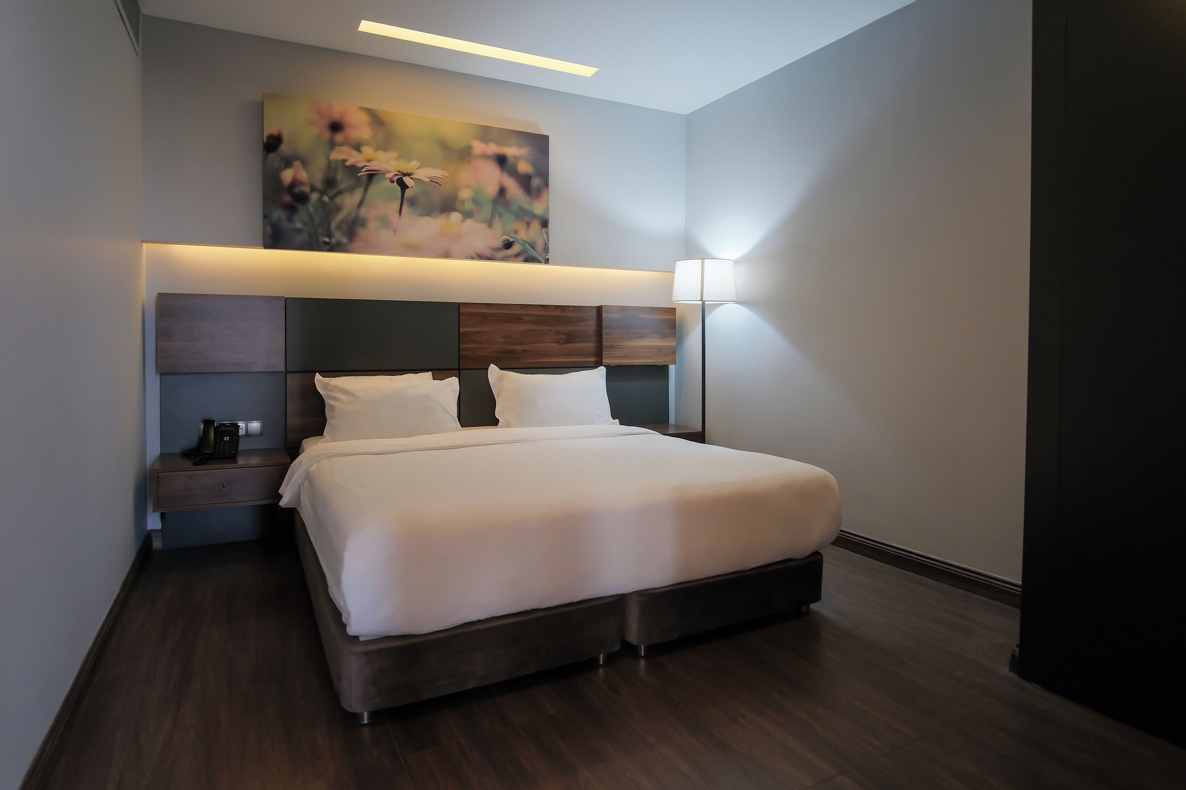 A modern hotel room with a large bed, a bedside table with a phone, a wall-mounted painting, and a floor lamp.