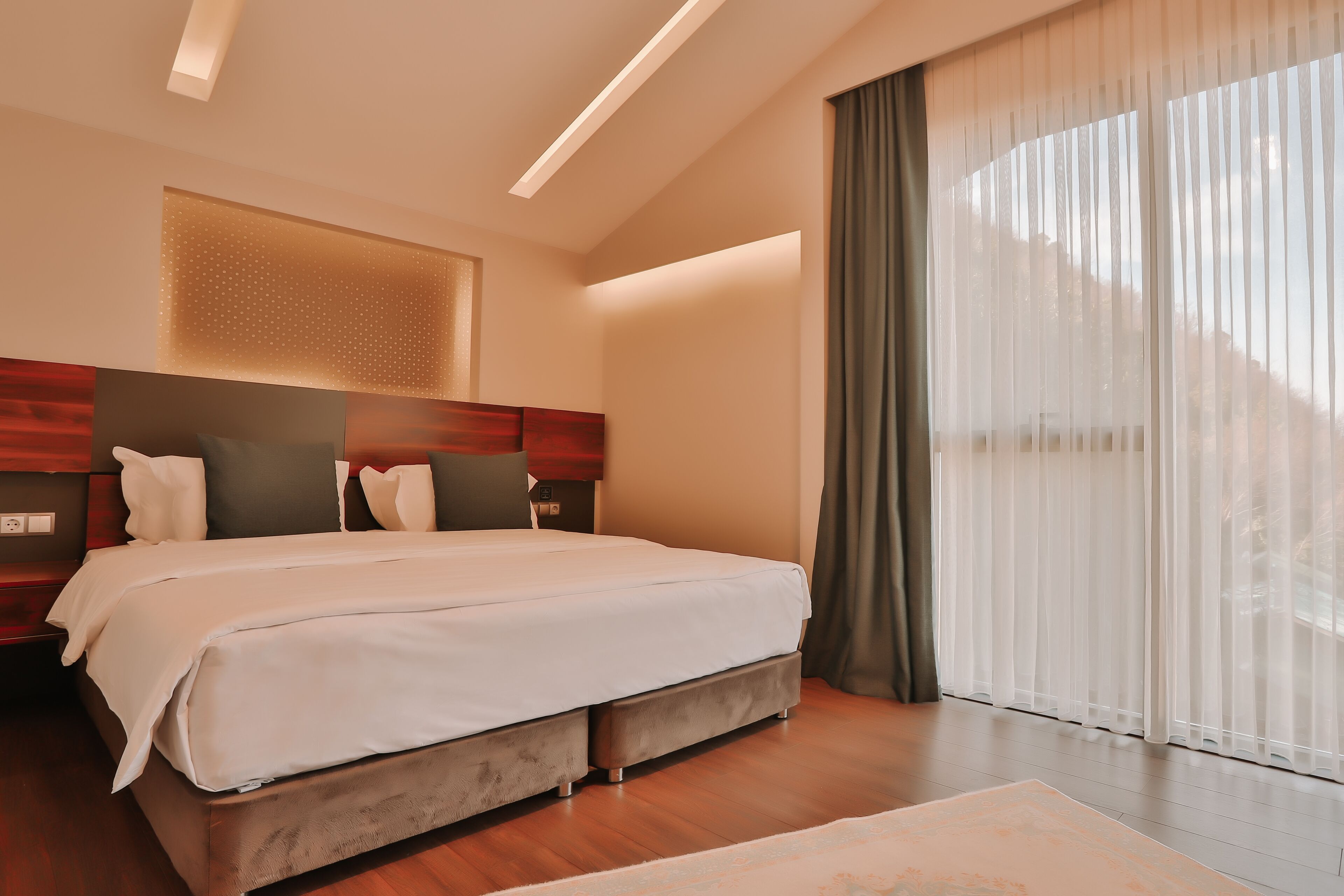 Un dormitori d'hotel modern amb un llit gran, un panell de fusta al capçal i una finestra ampla amb cortines de tela fina.