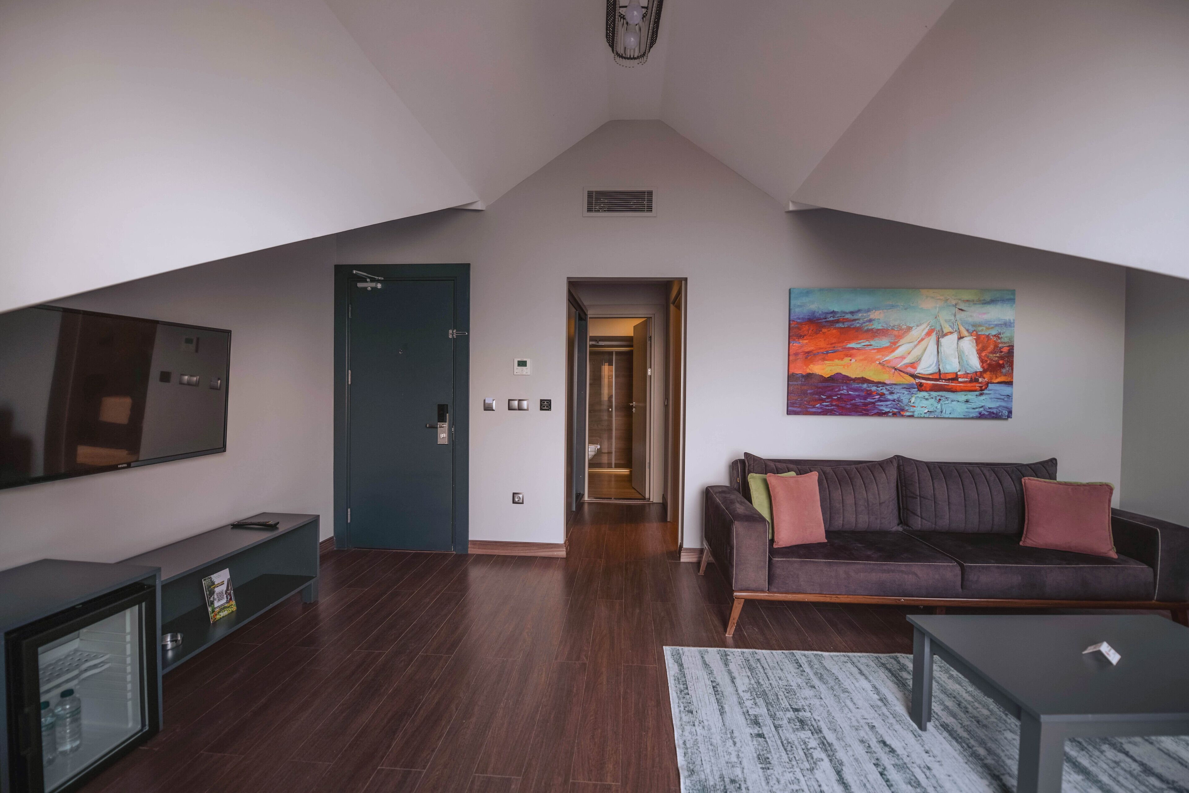 Un salon moderne avec un parquet en bois brun foncé, un canapé brun foncé agrémenté de coussins rouges, un téléviseur à écran plat posé sur un meuble noir et un tableau représentant un voilier accroché au mur.