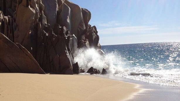 A beach with a rocky cliff and waves crashing onto the shore. - Fabulous Developers Beachfront PENTHOUSE XMAS & NY WKS @ Playa Grande Resort (Cabo San Lucas)