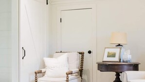 Un fauteuil blanc avec un coussin portant l’inscription « HAMHOUSE », dans une pièce où se trouvent une porte, une petite table avec une lampe et un cadre photo.