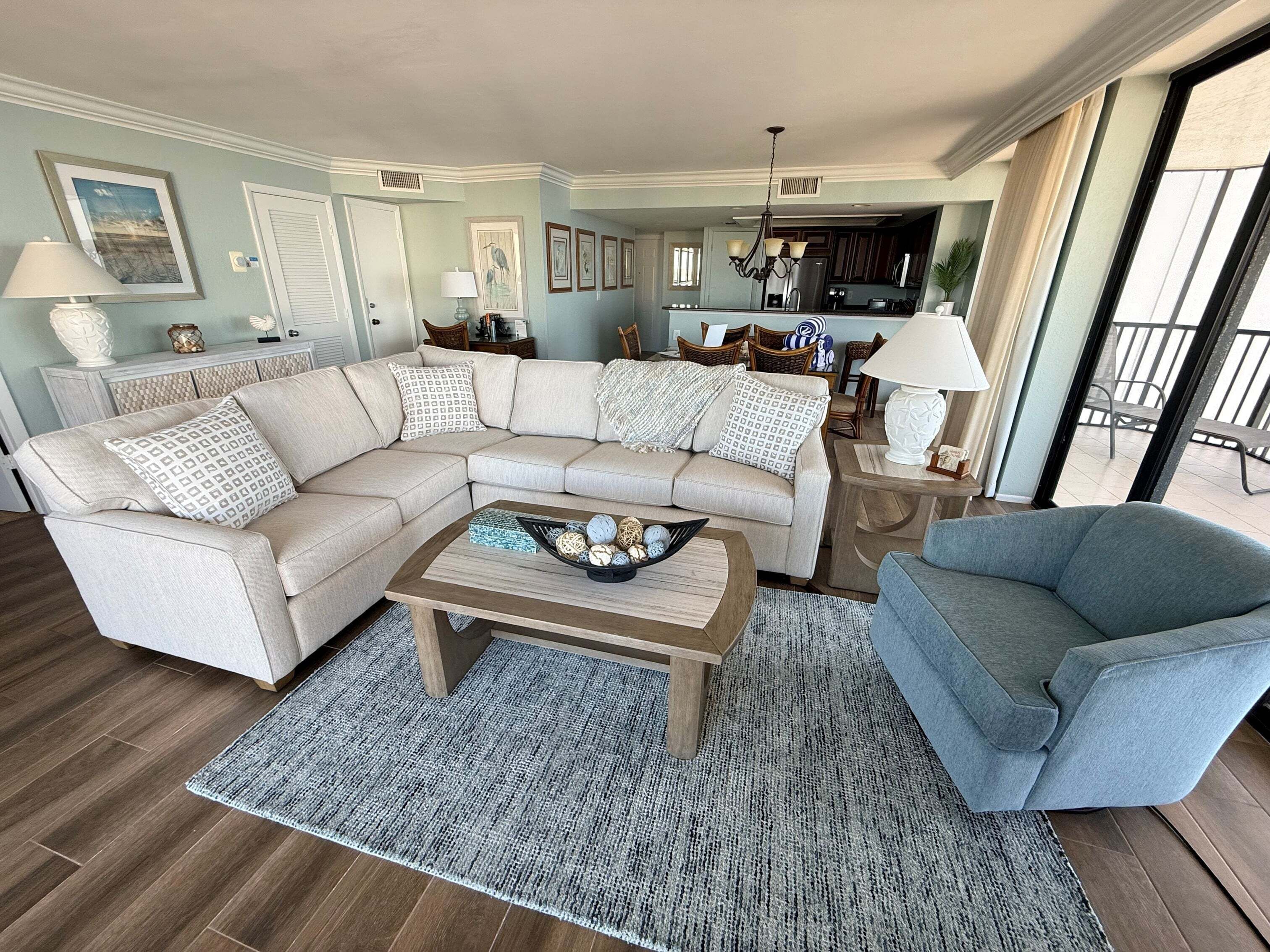 Una sala de estar con un sofá modular, una mesa de centro con objetos decorativos, un sillón azul y una cocina al fondo.
