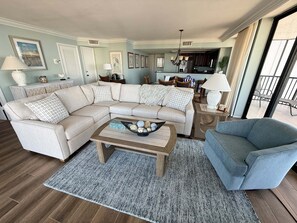 Ein Wohnzimmer mit einem Ecksofa, einem Couchtisch mit Dekoration, einem blauen Sessel und einer Küche im Hintergrund.