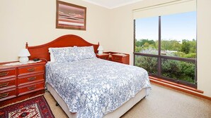 Un dormitorio con un ventanal grande, una cama con colcha estampada, una mesita de noche, un aparador y un cuadro en la pared.