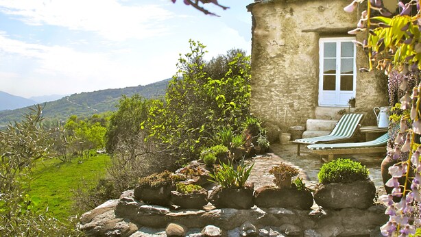 Une maison en pierre avec une porte et des fenêtres blanches, un chemin de pierre menant à une chaise et une table, et une vue sur des collines verdoyantes en arrière-plan.