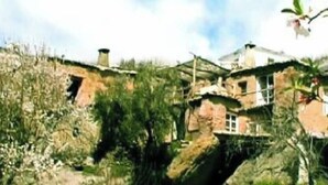 Un bâtiment rustique en pierre, à plusieurs niveaux, entouré d’une végétation luxuriante et sous un ciel dégagé.