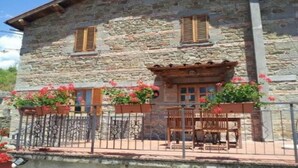 Un edificio de piedra con contraventanas de madera, un balcón con barandillas metálicas y jardineras con flores rosadas.
