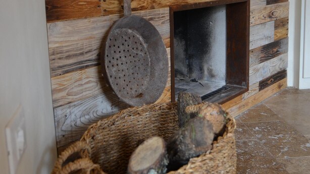 Een rustieke houten wand met een open haard, een hangende pannendrager en een rieten mand vol houtblokken.