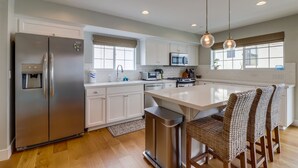 A modern kitchen with white cabinetry, stainless steel appliances, and a central island with wicker chairs. - Seaglass Shores: New 2BR/2BTH Modern Condo With Bay & Ocean Views (San Diego)