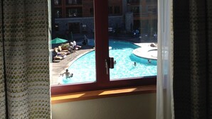A view from a hotel room window overlooking a swimming pool area with a building in the background.