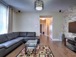 A modern living room with a sectional sofa, a glass coffee table, a patterned rug, a stone-textured fireplace, and a flat-screen TV.