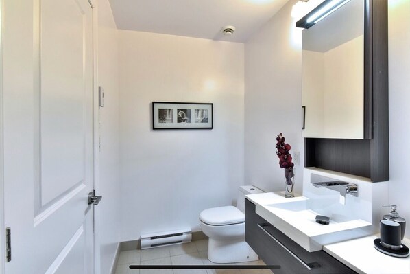 Casa de banho moderna com paredes brancas, sanita branca, toucador com espelho e quadro emoldurado na parede.