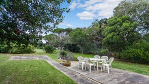 A patio area with a table and chairs, surrounded by lush greenery and trees. - Aireys Inlet Cottage (Aireys Inlet)