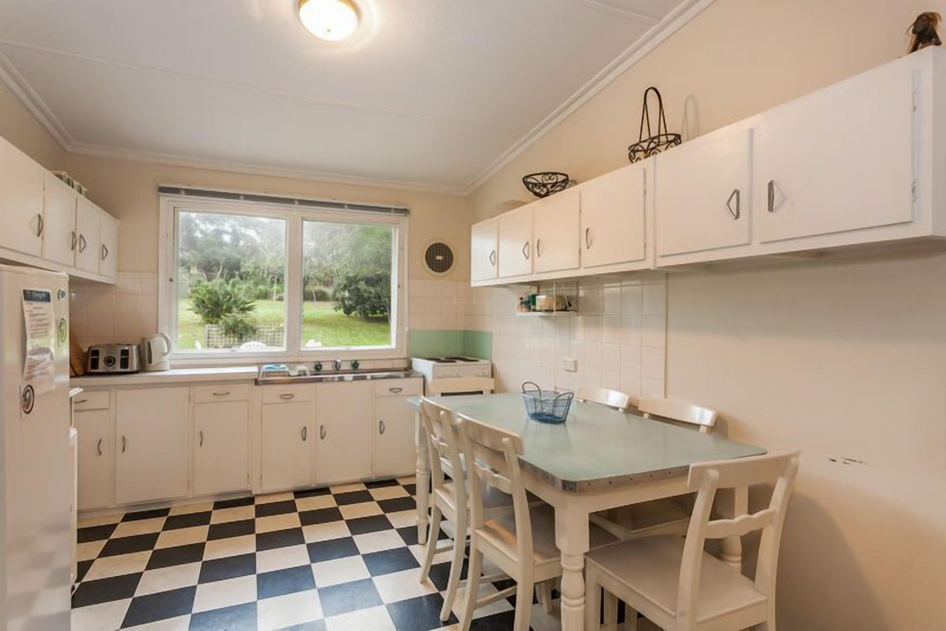 A kitchen with white cabinets, a checkered floor, a window with a view of greenery, and a table with chairs.