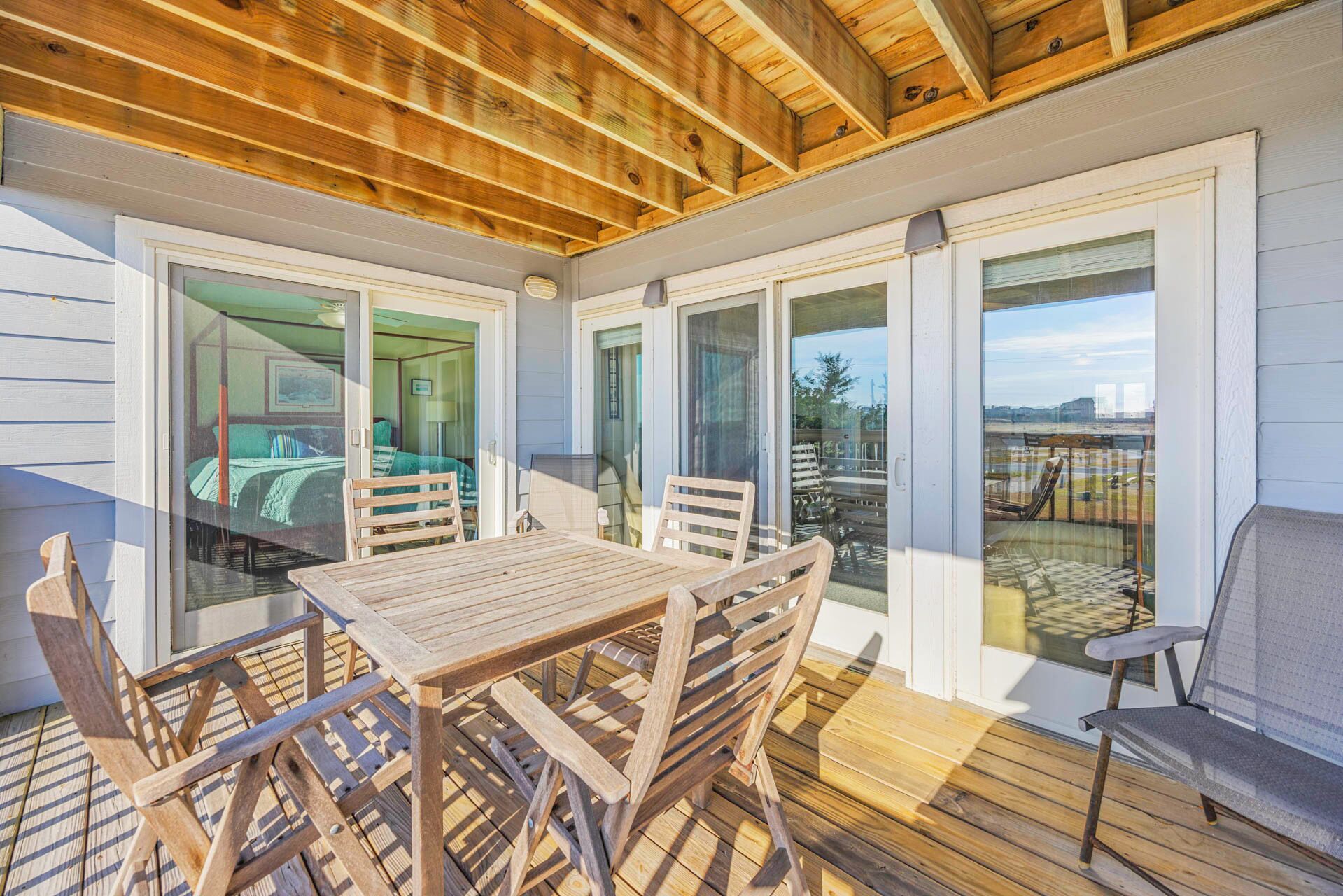 A covered patio with a wooden table and chairs, glass doors leading to a bedroom, and a view of the beach.