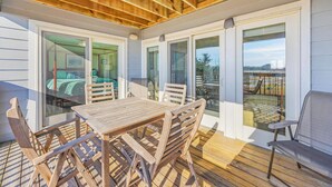Un patio cubierto con una mesa y sillas de madera, puertas de cristal que dan a un dormitorio y vista a la playa.