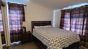 Une chambre à coucher avec un lit, un plancher en bois, des rideaux, une table de chevet et une lampe.