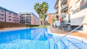 A swimming pool with clear blue water, surrounded by a tiled deck. There are palm trees and a building with balconies in the background.