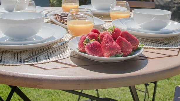 Ein Essplatz im Freien mit weißen Tellern, Schüsseln und Gläsern, einer Schale Erdbeeren und einem Glas Orangensaft.