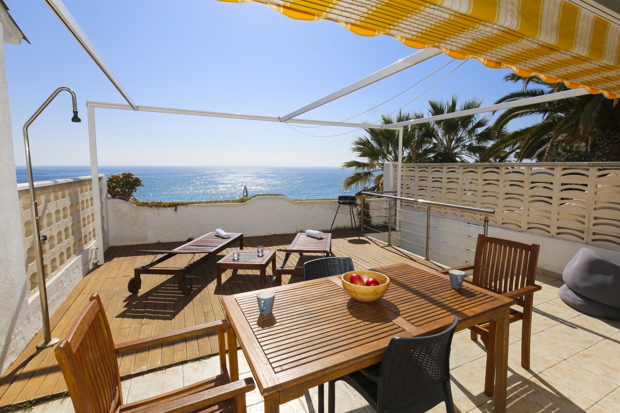 Una terrazza esterna con mobili in legno, un tavolo con una ciotola di frutta e vista sul mare.