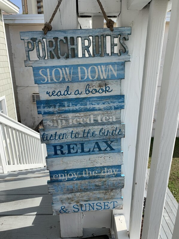 A wooden sign with the text 'PORCH RULES' and 'SLOW DOWN read a book listen to the birds RELAX enjoy the day & SUNSET'.