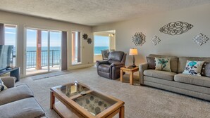 A spacious living room with a large window overlooking the ocean, a leather chair, a sofa with floral cushions, and decorative wall art.
