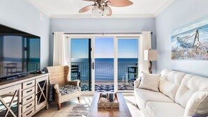 Un salon comprenant une télévision, un canapé, une table basse et un fauteuil. Il y a un balcon avec vue sur l’océan.