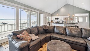 A modern living room with a large sectional sofa, a round ottoman, and a view of a body of water through large windows. - Crystal Lakefront Luxury Condo – North Shore, Crystal, Michigan (Crystal)