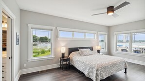 A bedroom with a large bed, two bedside tables, a ceiling fan, and multiple windows offering views of a body of water and greenery. - Crystal Lakefront Luxury Condo – North Shore, Crystal, Michigan (Crystal)