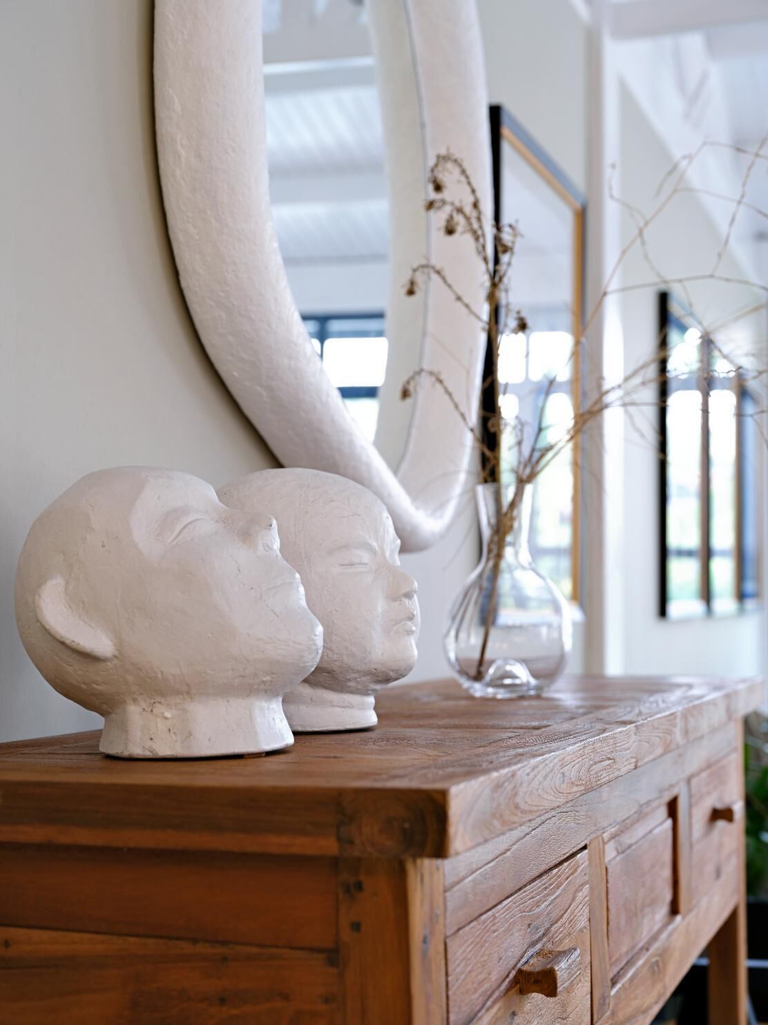 A wooden dresser with a large mirror, two white ceramic busts, and a vase with dried branches.
