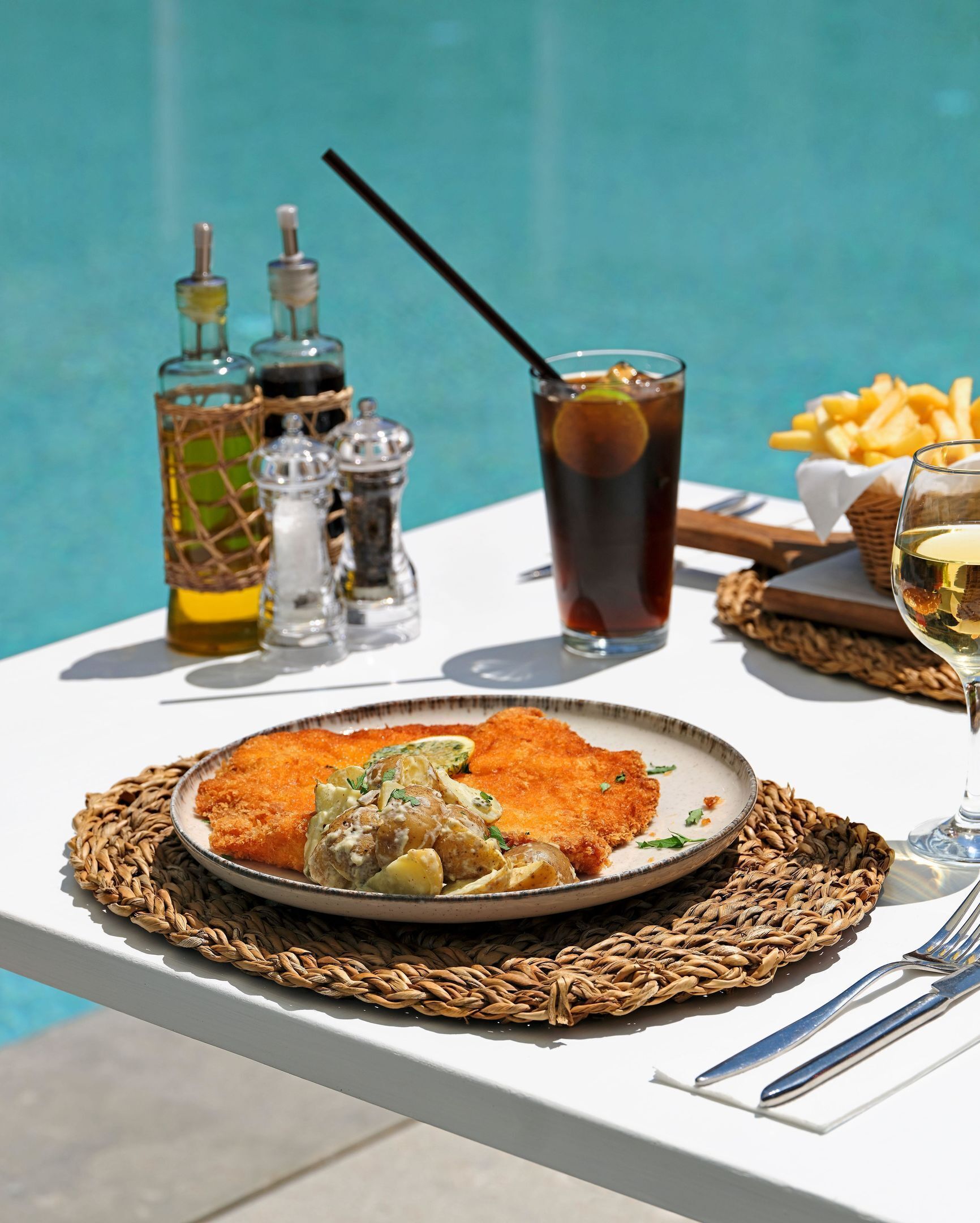 Una mesa con milanesa, papas, salsa, un vaso de bebida helada y una cesta de papas fritas junto a una piscina.