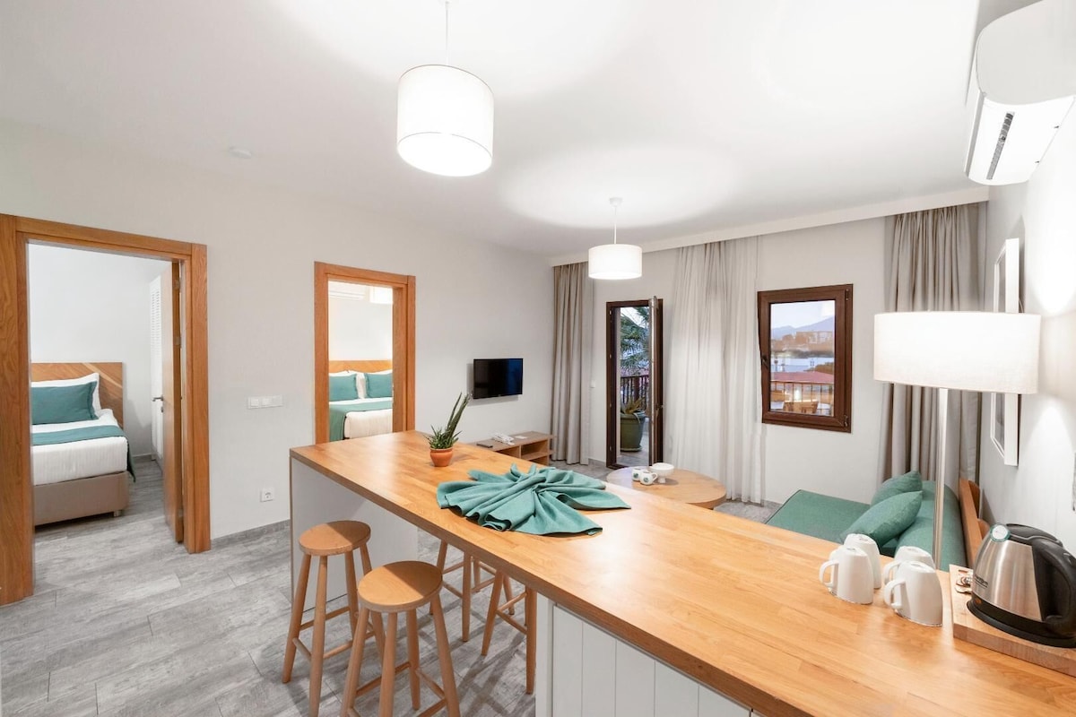A modern hotel room with a wooden dining area, a bed with teal bedding, a TV, and a view of the outdoors.
